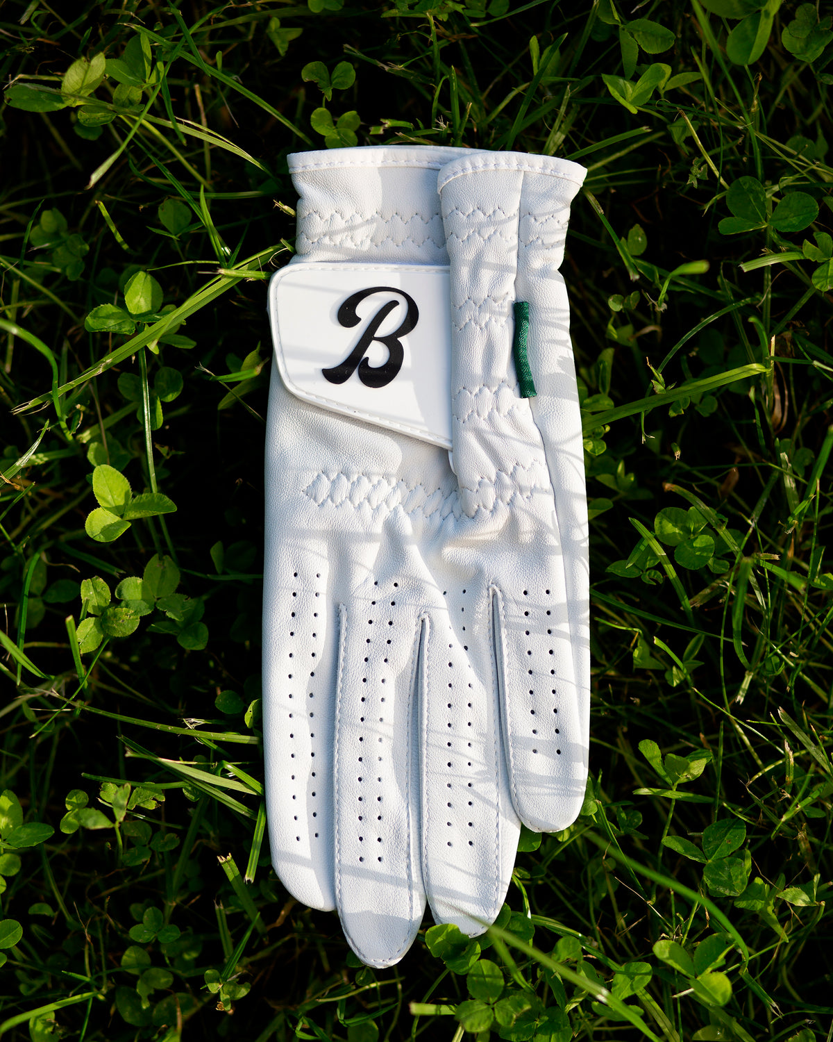 White Member Glove laid flat on grass, showing the black "B" logo patch on the back of the hand