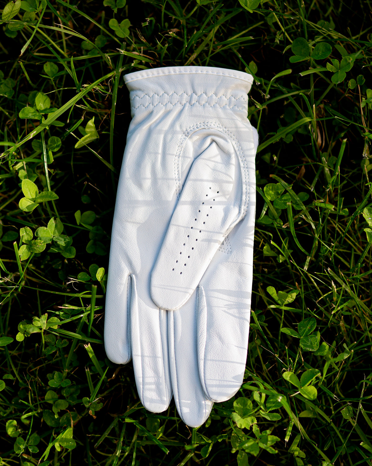 White Member Glove laid palm-down on grass, showing the leather palm and stitching