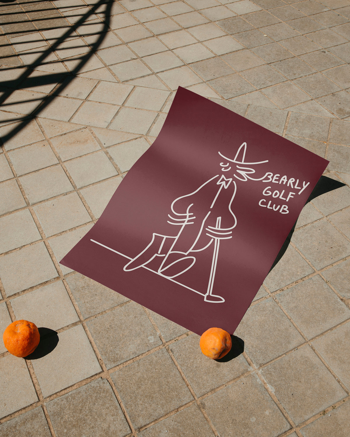 Maroon 50x70cm art print laid outdoors on paving stones, corners weighted with oranges