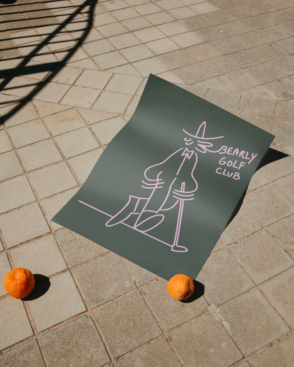 Dark green art print laid outdoors on paving stones, corners weighted with oranges