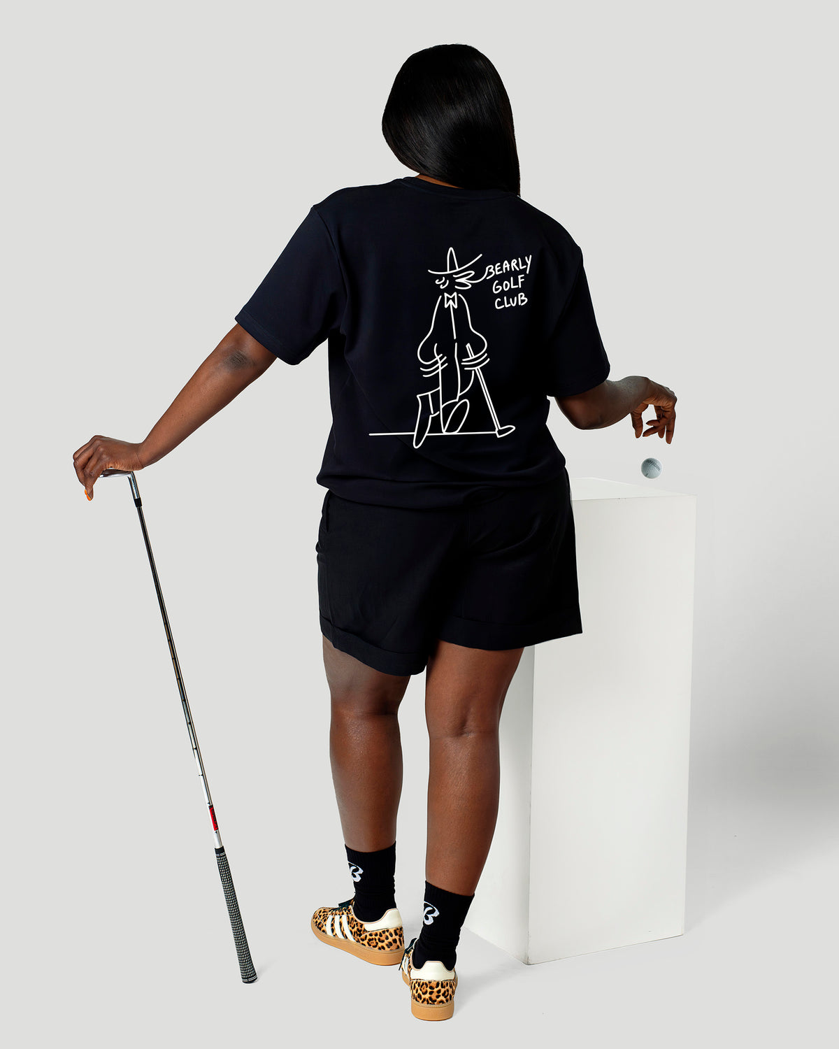 Woman seen from behind in the black Gus Gus Tee, white character artwork on the back, holding a golf club